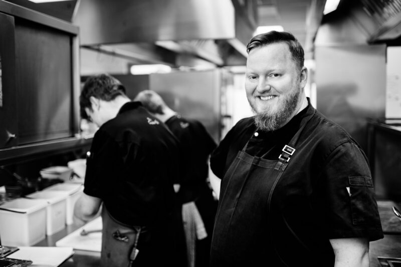 Smiling chef Dominik Obermeier stands in a kitchen, with two colleagues working in the background.
