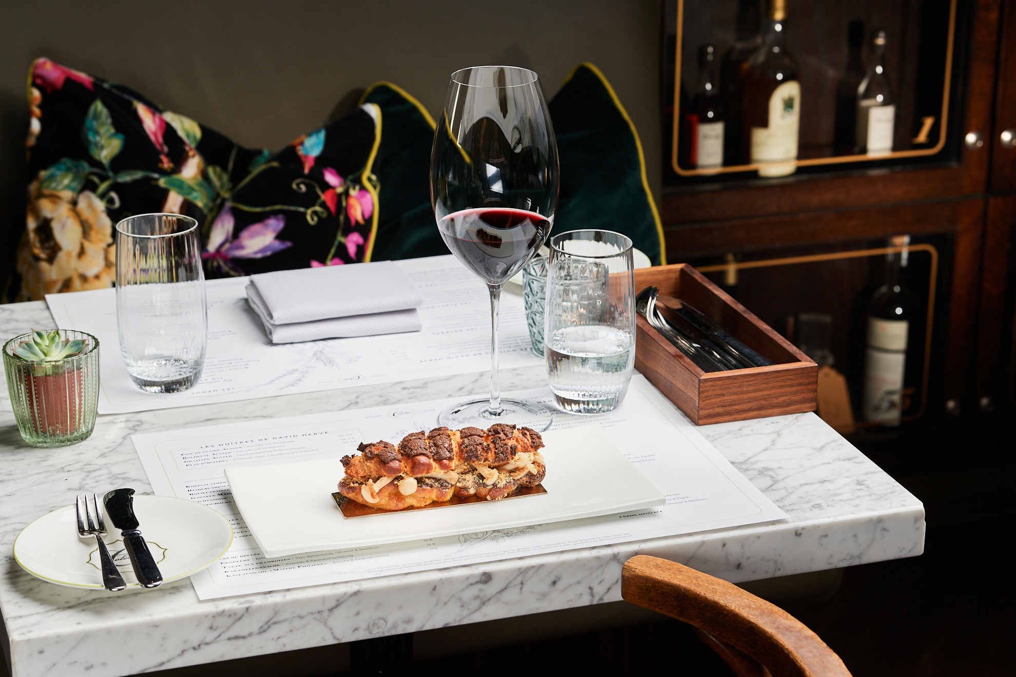 Eclair on a white plate, accompanied by a glass of red wine, set on a marble table with decorative cushions in the backgro...