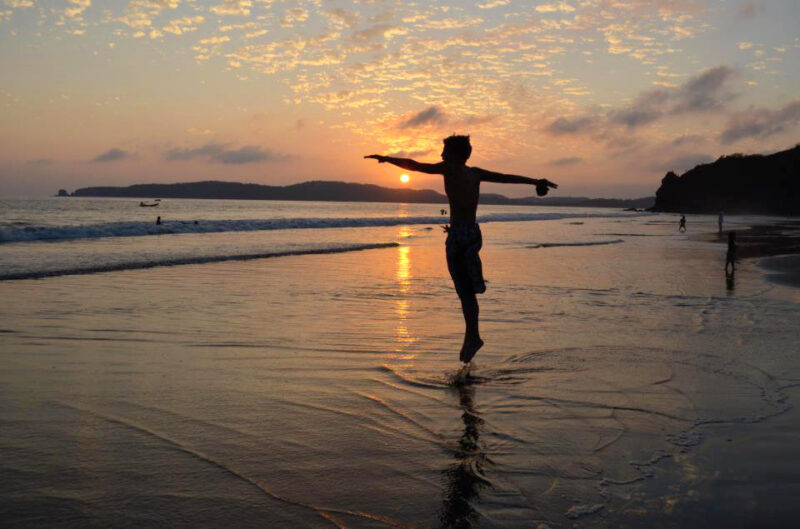 Silhouette of a person jumping on a beach at sunset, with waves and clouds in the background.
