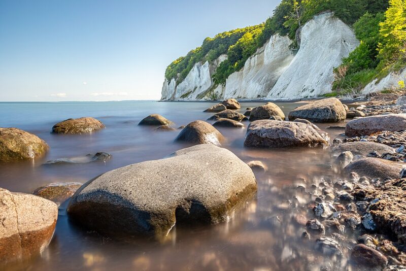 Rocky shoreline with smooth boulders in foreground, leading to white chalk cliffs and lush greenery under a clear blue sky.