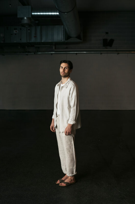 Man in a light-colored outfit stands in an empty, industrial space with a neutral background.