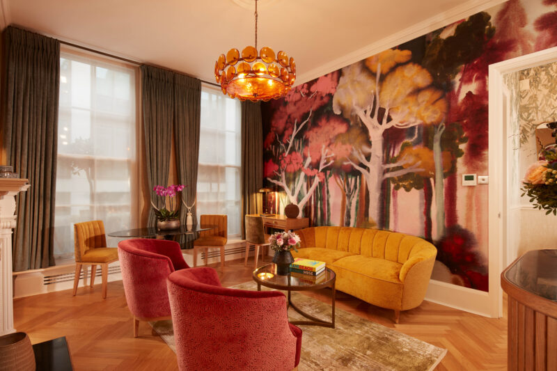 Colorful living room featuring a yellow sofa, red chairs, a round table, and a vibrant forest mural on the wall.
