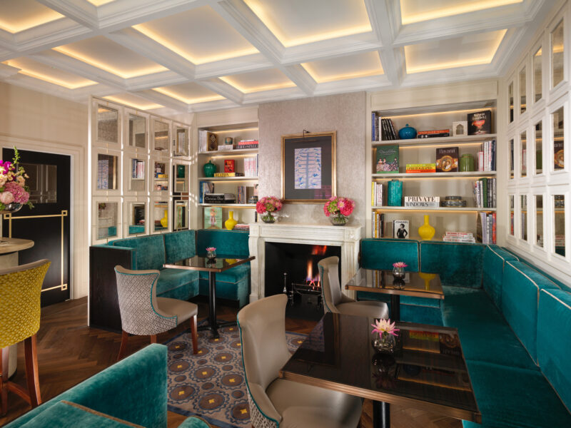 Elegant drawing room at Flemings Mayfair, featuring teal seating, a fireplace, and bookshelves with decorative items.