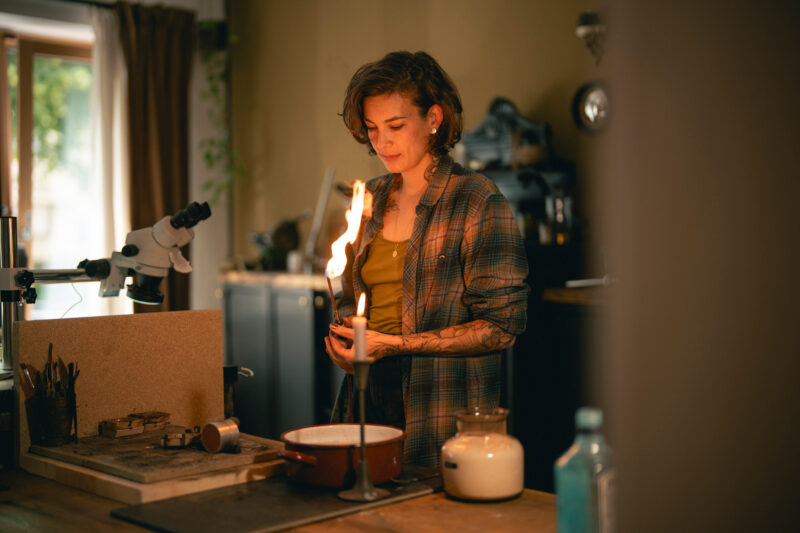 Woman with short hair and tattoos holding a flame in a workshop, surrounded by tools and equipment.