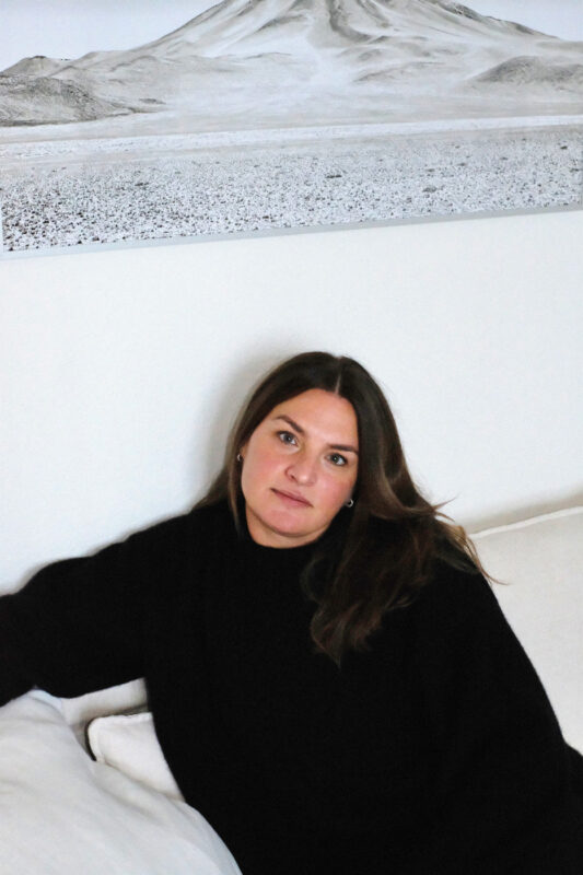 Woman with long brown hair wearing a black sweater, sitting on a couch, with a mountain landscape artwork in the background.