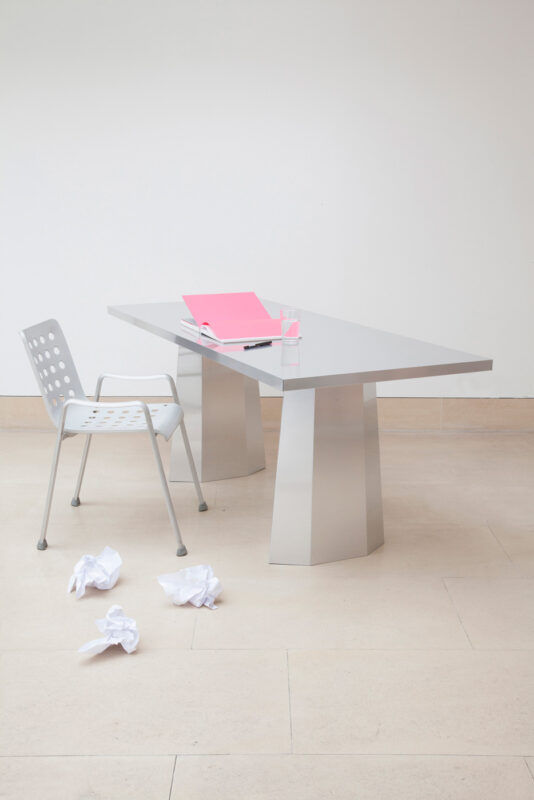 Modern silver table with two geometric legs, a pink notebook, and a chair on a light stone floor, crumpled paper nearby.