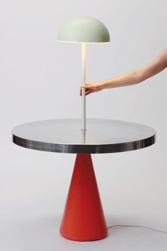 Modern table with a conical orange base and a circular metallic top, featuring a hand adjusting a green lamp.
