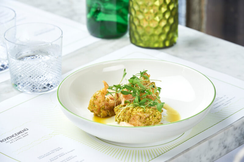 Three Moroccan-style shrimp garnished with herbs, served in a white bowl on a marble table, with drinks in the background.