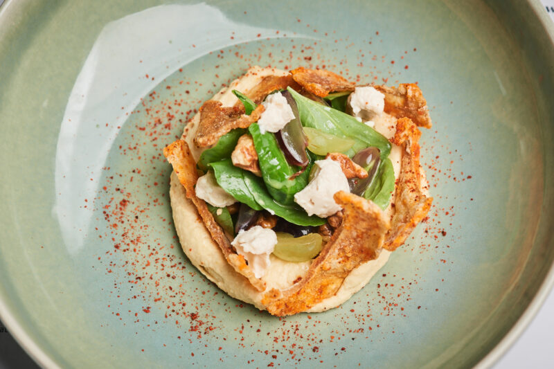 Hummus topped with greens, olives, and crispy chips, served in a light green bowl with a sprinkle of paprika.
