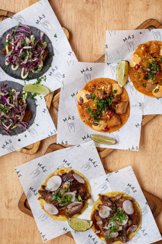 Five colorful tacos on branded paper, featuring various toppings like onions, cilantro, and lime wedges.