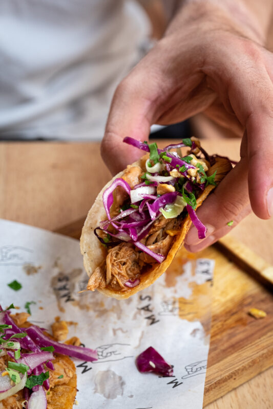 A hand holding a taco filled with shredded chicken, purple cabbage, and garnishes, with another taco on a wooden surface.