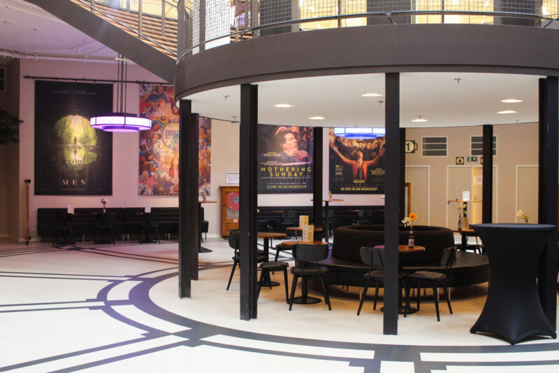 Modern interior of a lobby featuring circular seating, posters for "Mothering Sunday" and "Men," and a bar table.