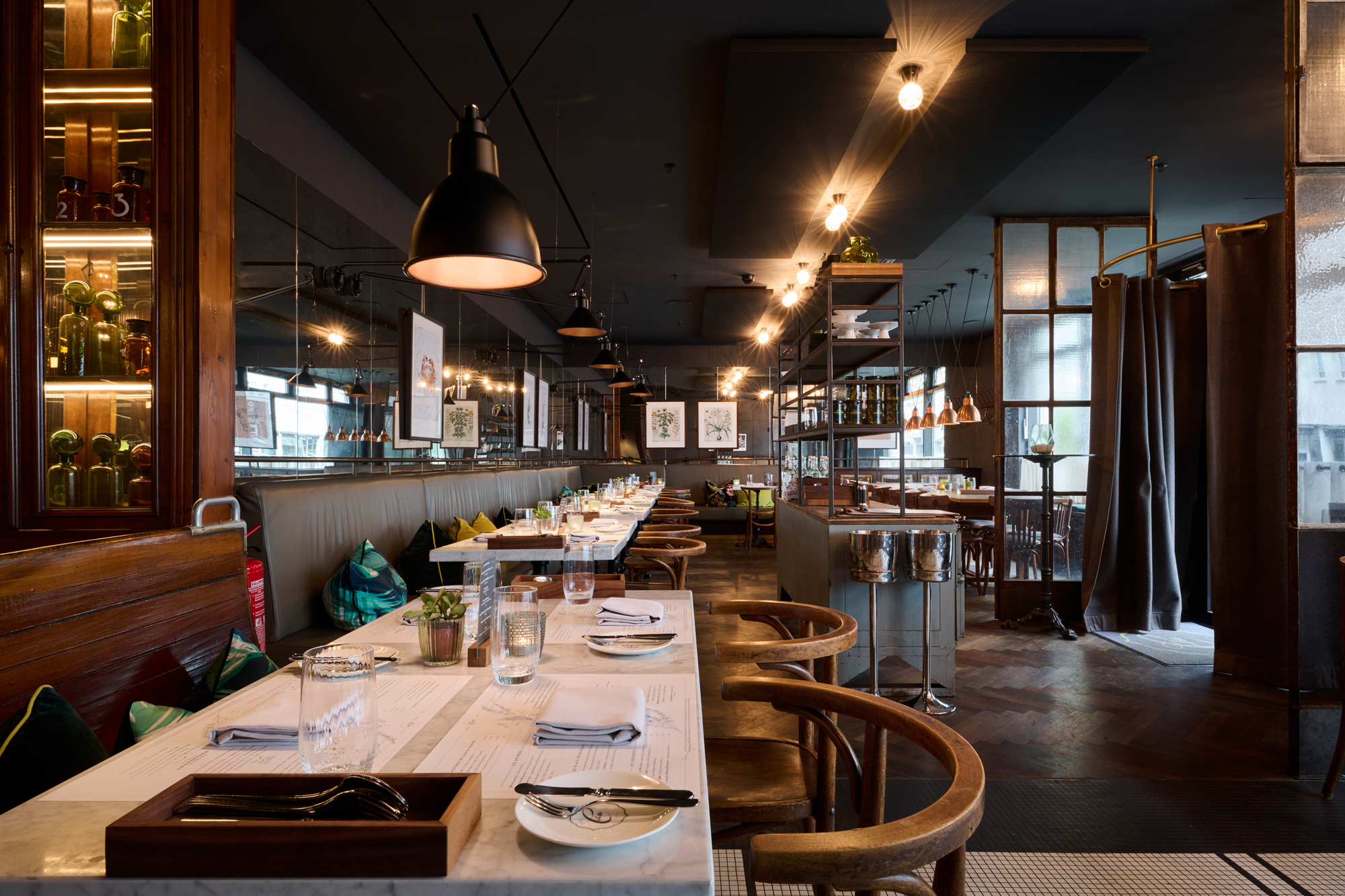 Modern restaurant interior featuring long tables, warm lighting, and stylish decor, with plants and artwork on the walls.