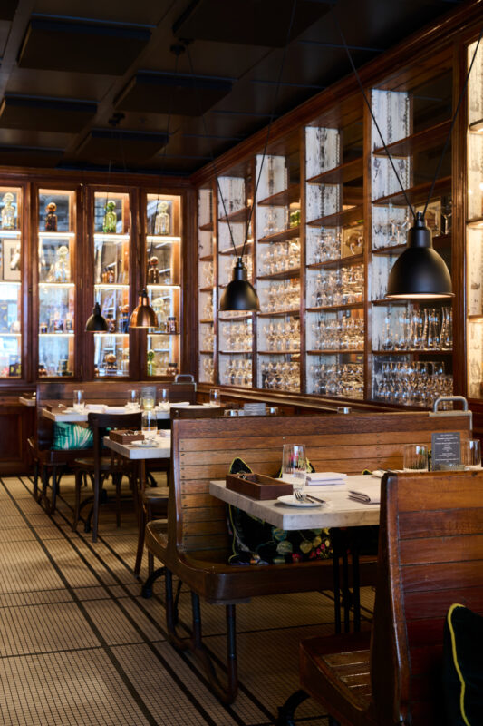Elegant restaurant interior with wooden benches, marble tables, and glassware displayed in illuminated cabinets.