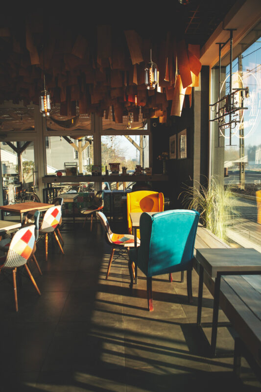 Colorful chairs and tables in a modern café with wooden ceiling decor and large windows letting in sunlight.