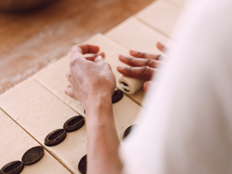 Hands rolling dough with chocolate discs on a wooden surface, part of a baking process.