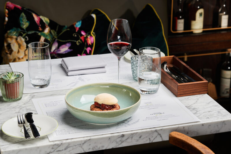 A dessert featuring a pastry with cream on a green plate, accompanied by a glass of red wine and water on a marble table.