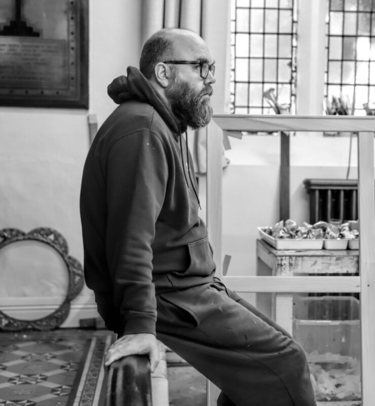 Nathan Walsh seated in a studio, wearing glasses and a hoodie, with art supplies and a workbench in the background.