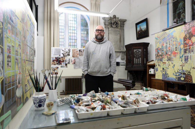 Artist Nathan Walsh stands in his studio, surrounded by paint tubes and artworks, with large windows in the background.