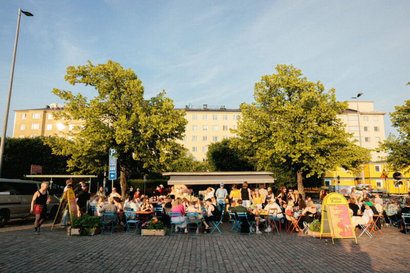 Busy outdoor café scene with people dining at blue tables under trees, vibrant buildings in the background.