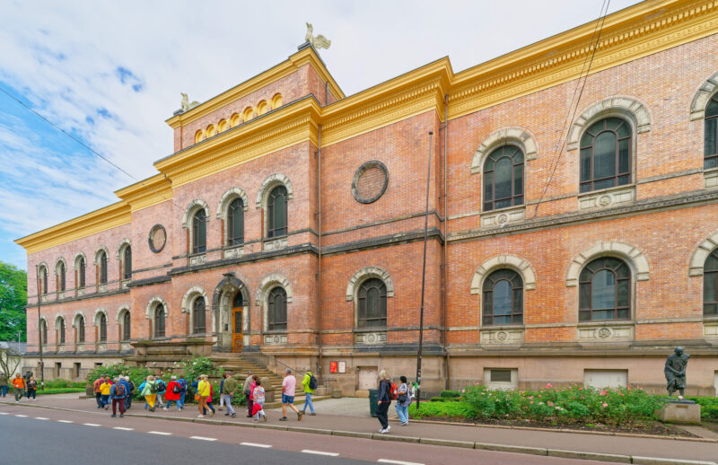 Brick building of the National Gallery in Oslo with pedestrians walking by, under a partly cloudy sky.