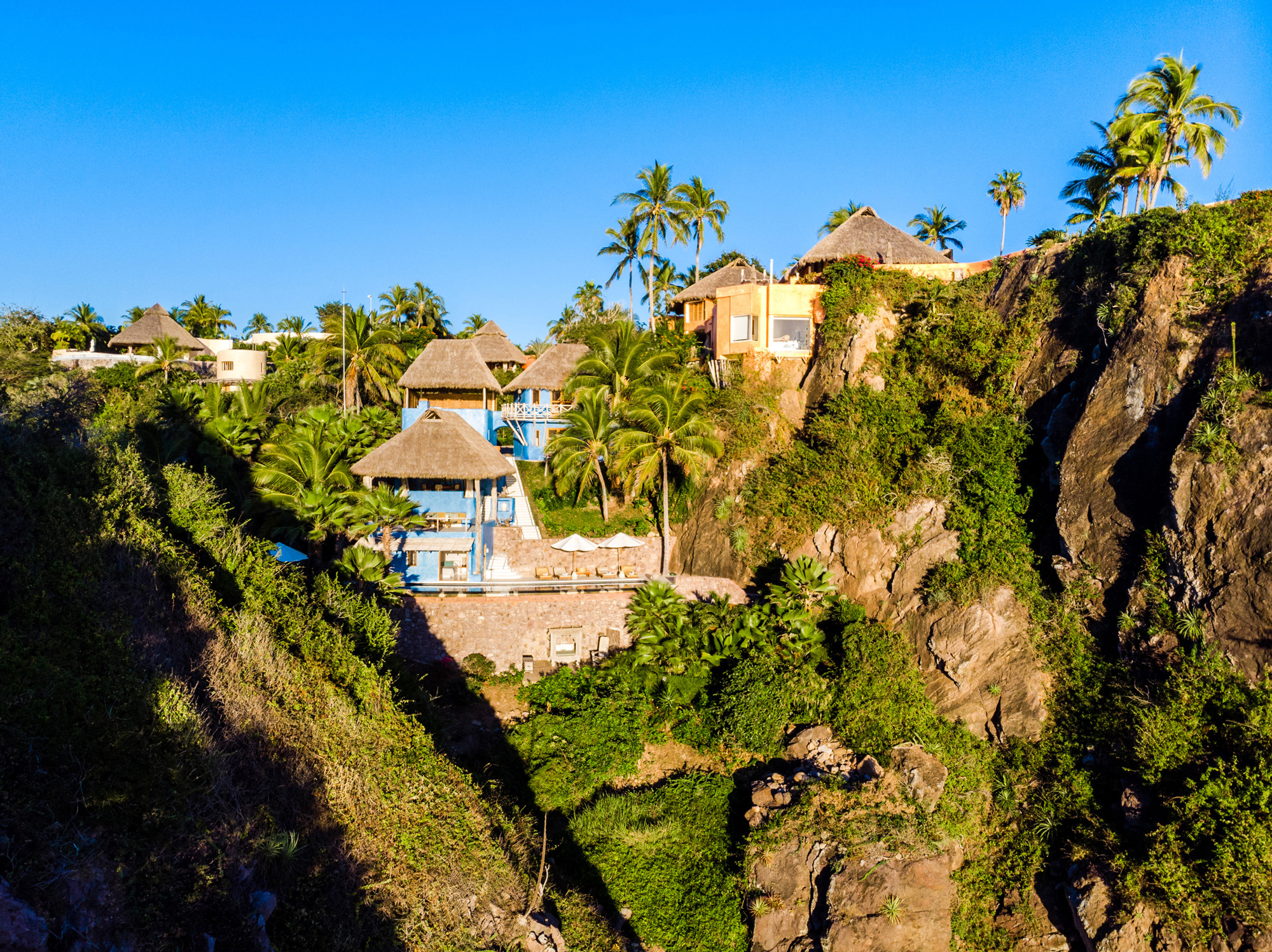 Tropical hillside with thatched-roof houses and palm trees under a clear blue sky, part of Nido de Amor resort.
