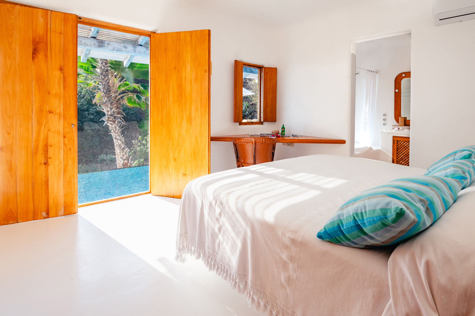 Bright bedroom in Nido de Amor with a white bed, wooden doors open to a garden, and a mirror on the wall.