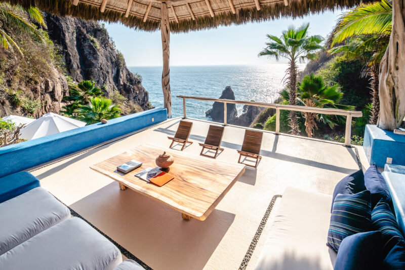Cozy outdoor lounge with a wooden table, two chairs, and ocean view, surrounded by palm trees and rocky cliffs.