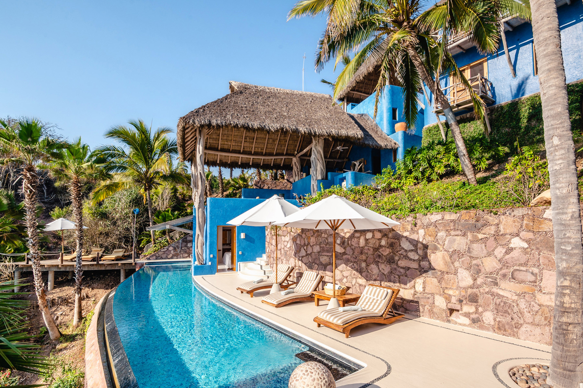 Luxury poolside area with loungers, palm trees, and a blue building under a thatched roof in a tropical setting.