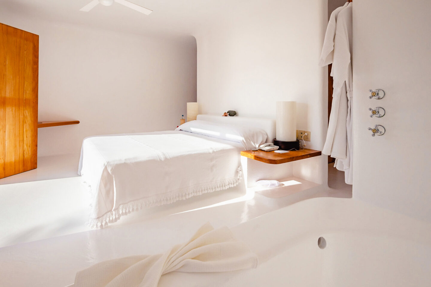 Bright, minimalist bedroom featuring a white bedspread, wooden accents, and a soaking tub, with soft natural light.