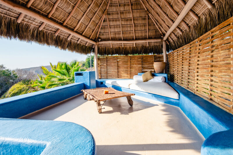 Open-air seating area with a thatched roof, blue walls, and a wooden table, surrounded by tropical greenery.