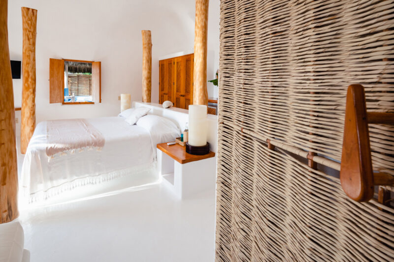 Bright, minimalist bedroom with a white bedspread, wooden pillars, and woven wall details, featuring a mirror and natural ...