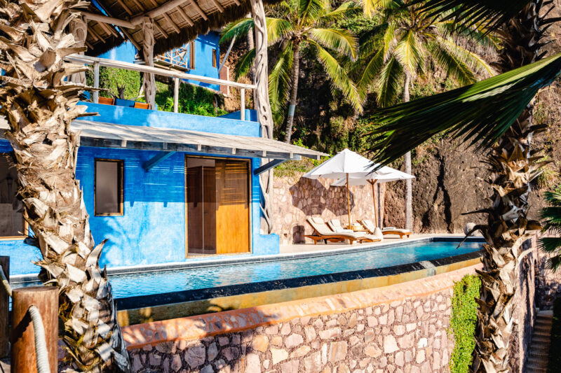 Vibrant blue building with a pool, surrounded by palm trees and lounge chairs, set against a rocky backdrop.