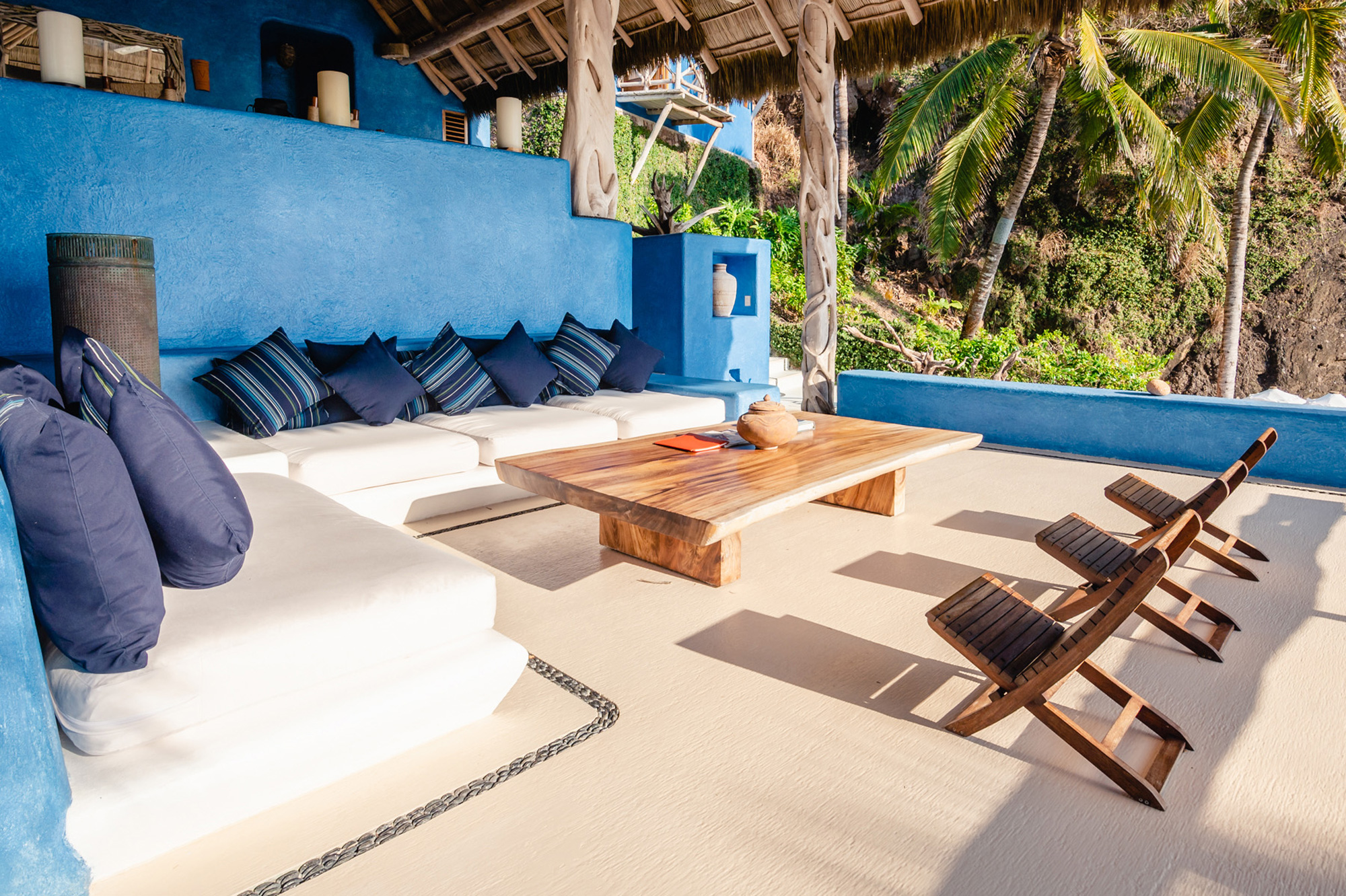 Lounge area with blue walls, a wooden table, and seating, surrounded by tropical greenery.