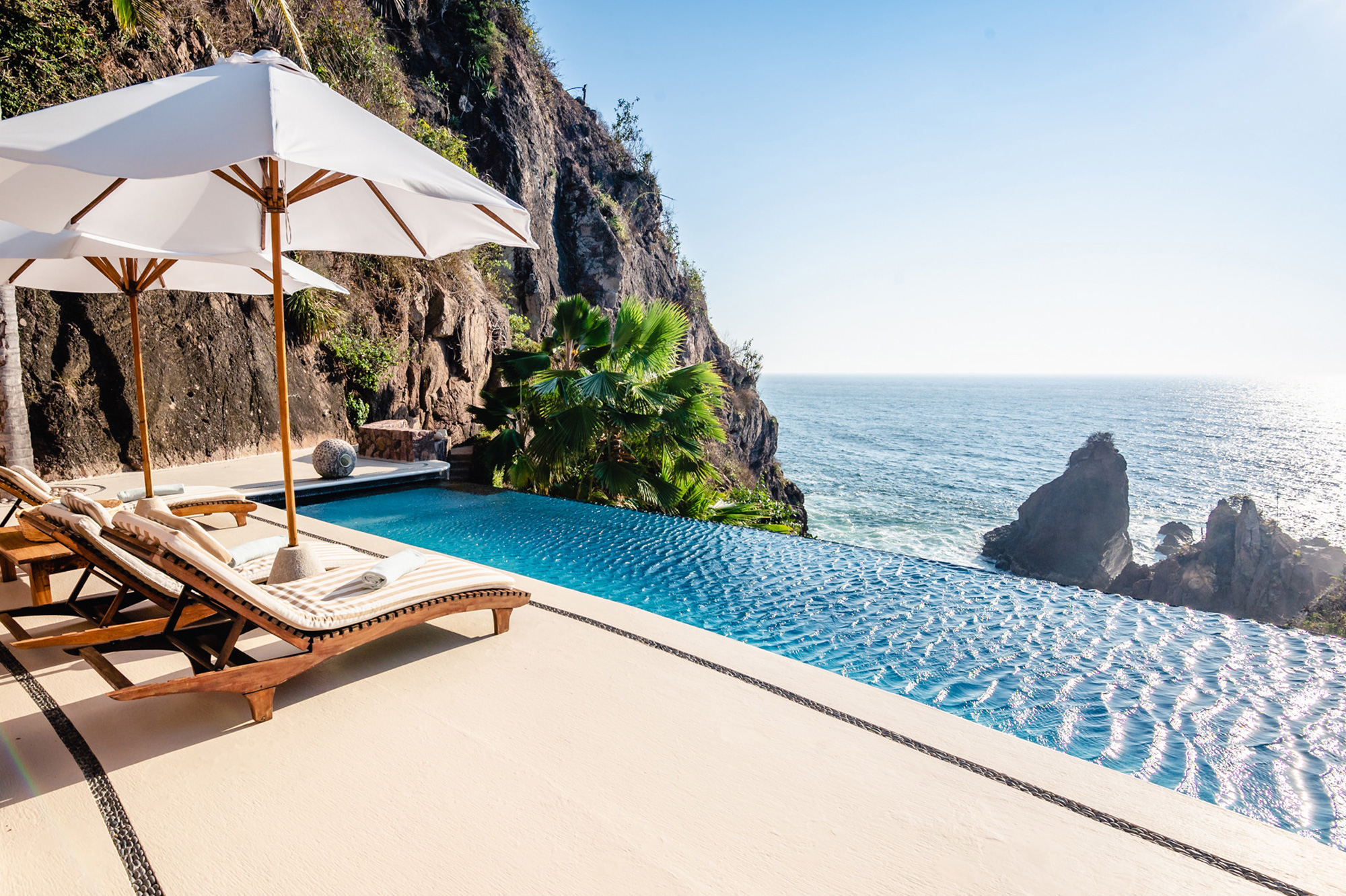 Infinity pool overlooking the ocean, with lounge chairs and umbrellas beside rocky cliffs under a clear blue sky.