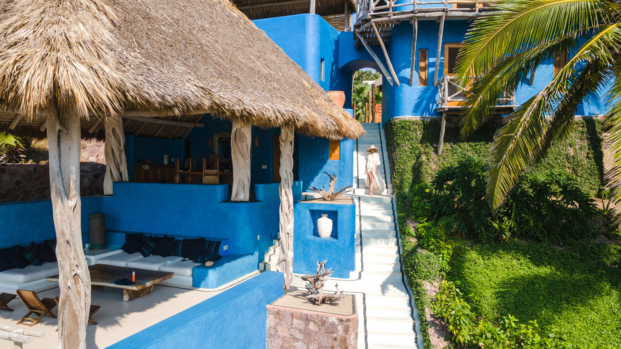 Tropical villa with a thatched roof, blue walls, and a staircase leading to a lush garden.