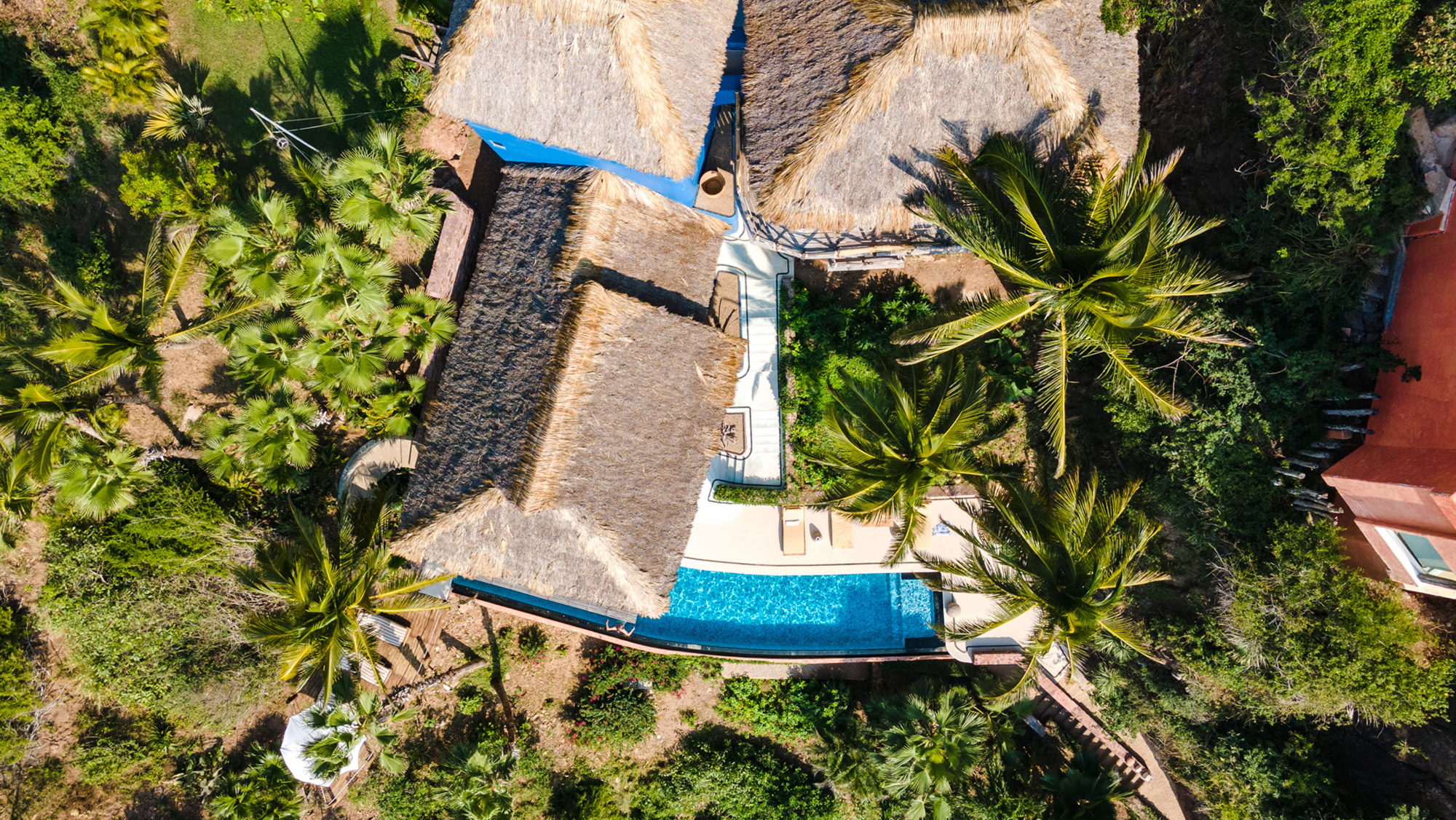 Aerial view of a tropical villa with thatched roofs, surrounded by palm trees and a swimming pool.