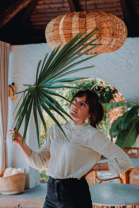 Evalie Wagner holding a large green leaf indoors, with a woven pendant light and plants in the background.