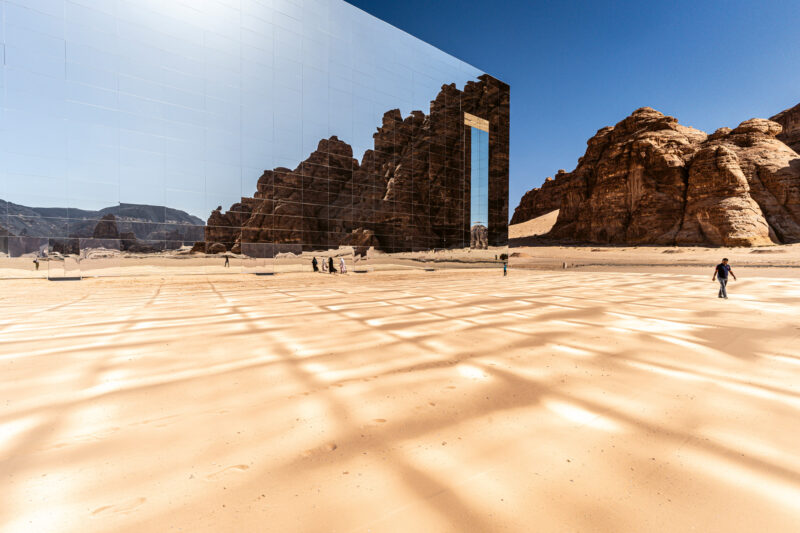 Reflective building against rocky landscape in Saudi Arabia, with people walking on sandy terrain under a clear blue sky.