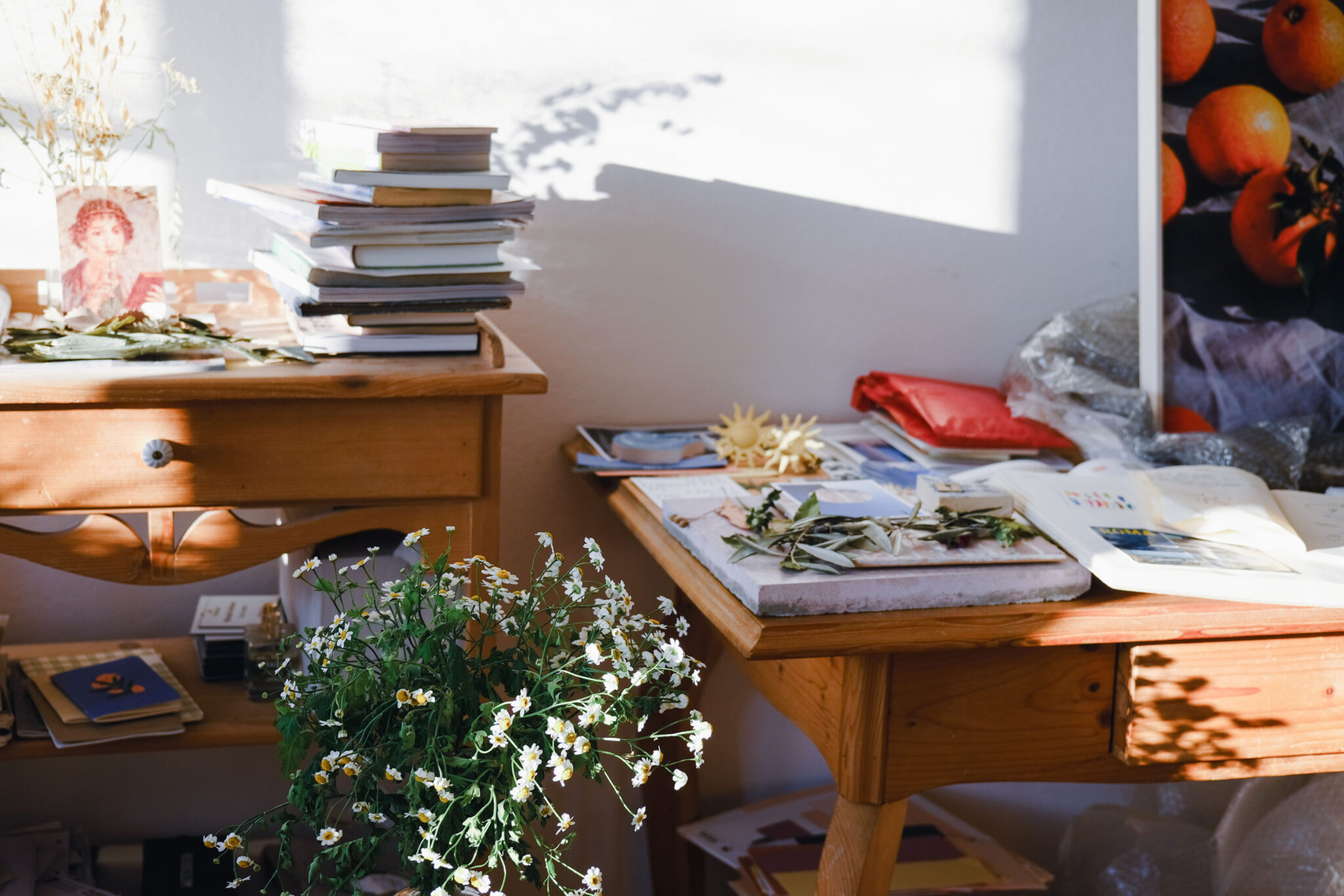 Sunlit studio space with wooden tables, stacked books, scattered papers, and a vase of flowers.