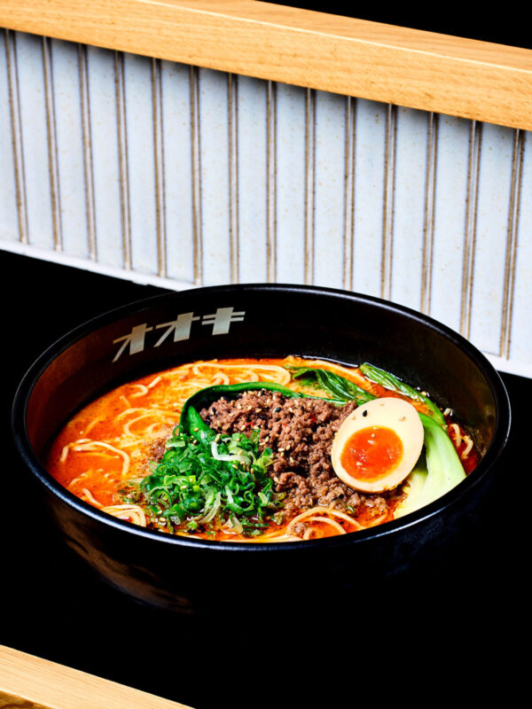 Bowl of tantanmen ramen with minced meat, soft-boiled egg, greens, and noodles against a wooden backdrop.