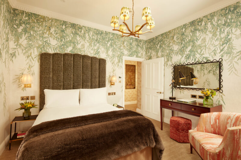 Elegant bedroom in a three-bedroom suite featuring a plush bed, floral wallpaper, a stylish mirror, and cozy seating.