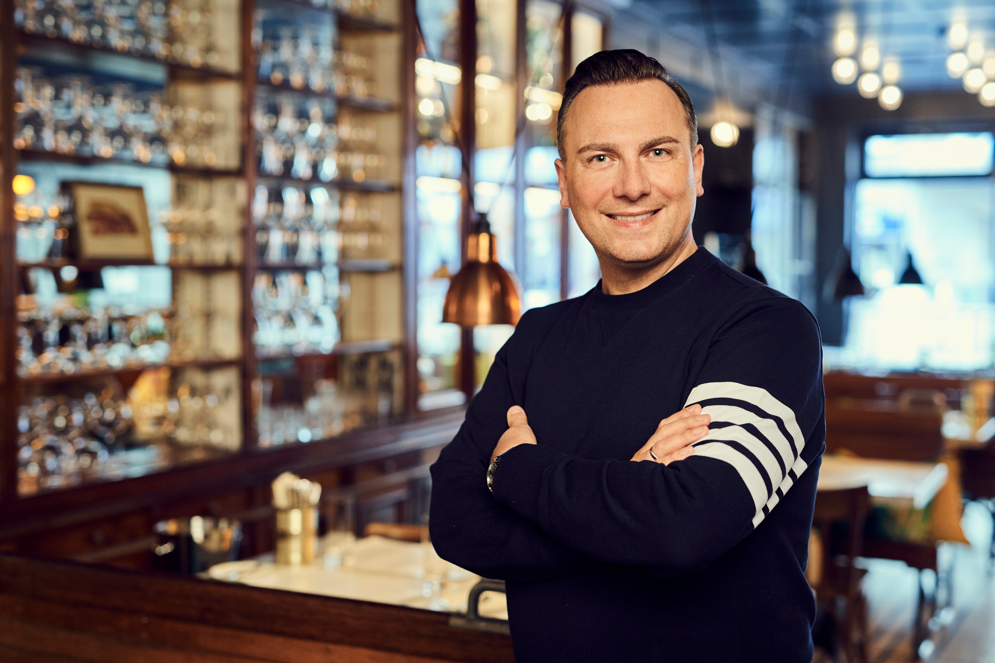 Tim Raue smiling with arms crossed, standing in Brasserie Colette, featuring a stylish interior with glassware in the back...