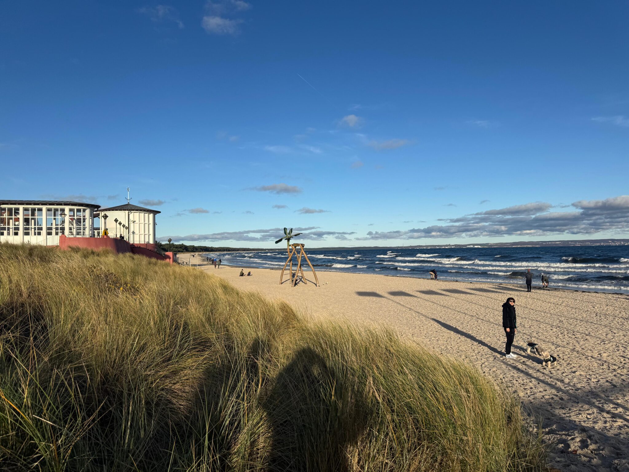 Beach scene at Kurhaus Binz with tall grass, a wooden structure, and people walking along the shore under a blue sky.