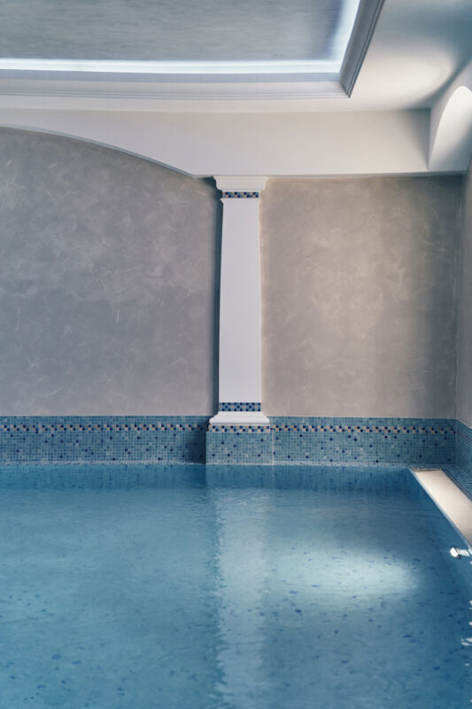 Well-lit indoor pool area featuring a textured gray wall, a decorative column, and a blue-tiled edge.