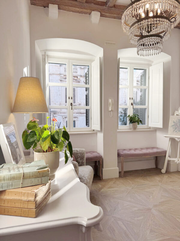 Bright interior of a room with two windows, a lamp, potted plant, and books on a table, showcasing a cozy atmosphere.