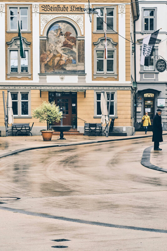 Empty street scene featuring a building with a mural, a person in a yellow coat, and a man in a suit.