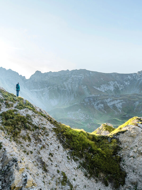 A person in a teal jacket stands on a rocky hillside, overlooking a mountainous landscape at sunrise.