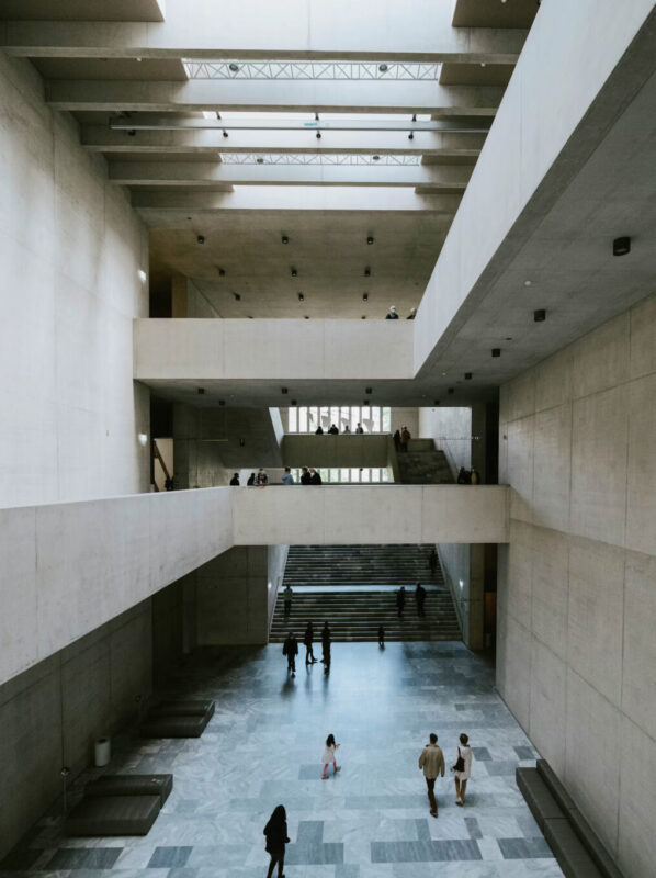 Modern concrete interior of a spacious building with multiple levels and people walking, illuminated by natural light.