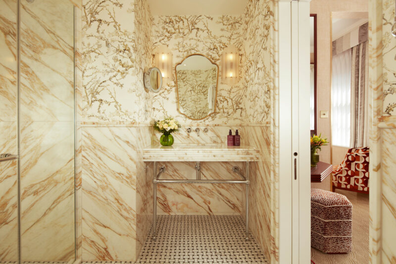 Luxurious bathroom with marble walls, a floral mirror, and a double sink, featuring a decorative vase of flowers.